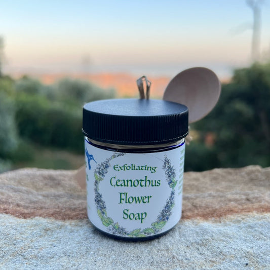 1.7 oz Cobalt blue glass jar of Ceanothus Flower Powder with wooden spoo for measuring on a rock with sunset of Refugio Gardens Ranch Sunset in the background.