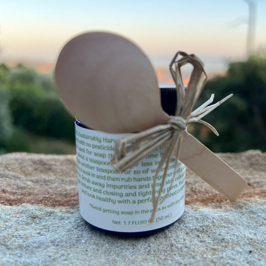 1.7 oz Cobalt blue glass jar of Ceanothus Flower Powder with wooden spoo for measuring on a rock with sunset of Refugio Gardens Ranch Sunset in the background.