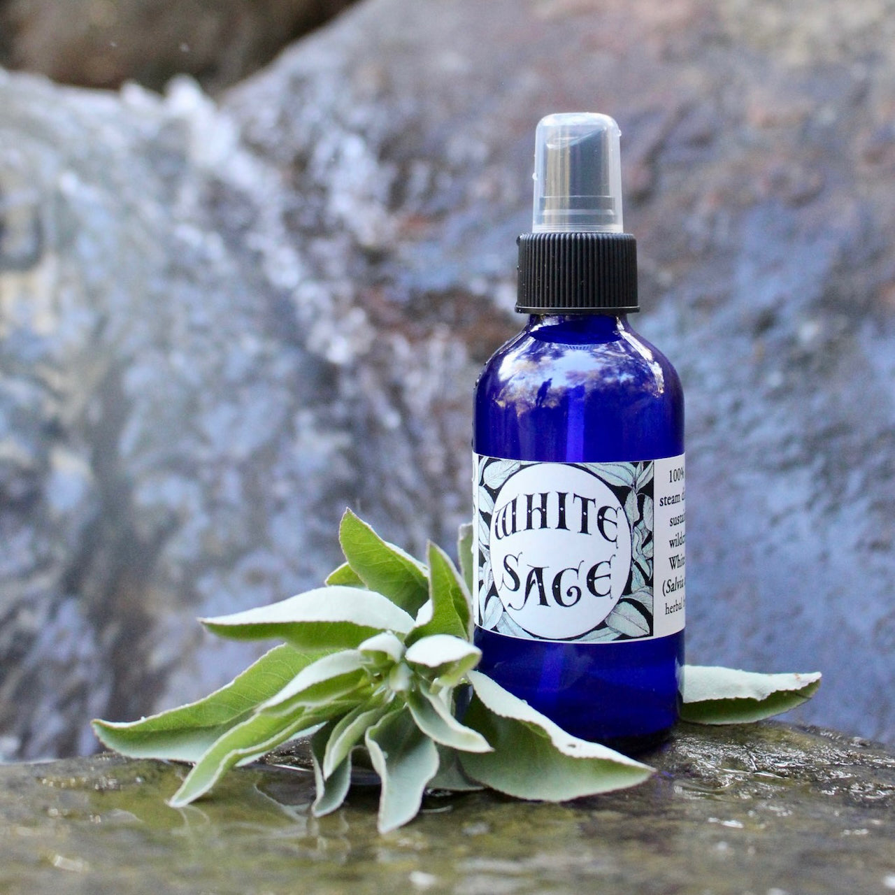 Photograph of a 4 oz cobalt blue glass spray bottle filled with White Sage hydrosol on rock withwhite sage leaves in a creek with a waterfall behind