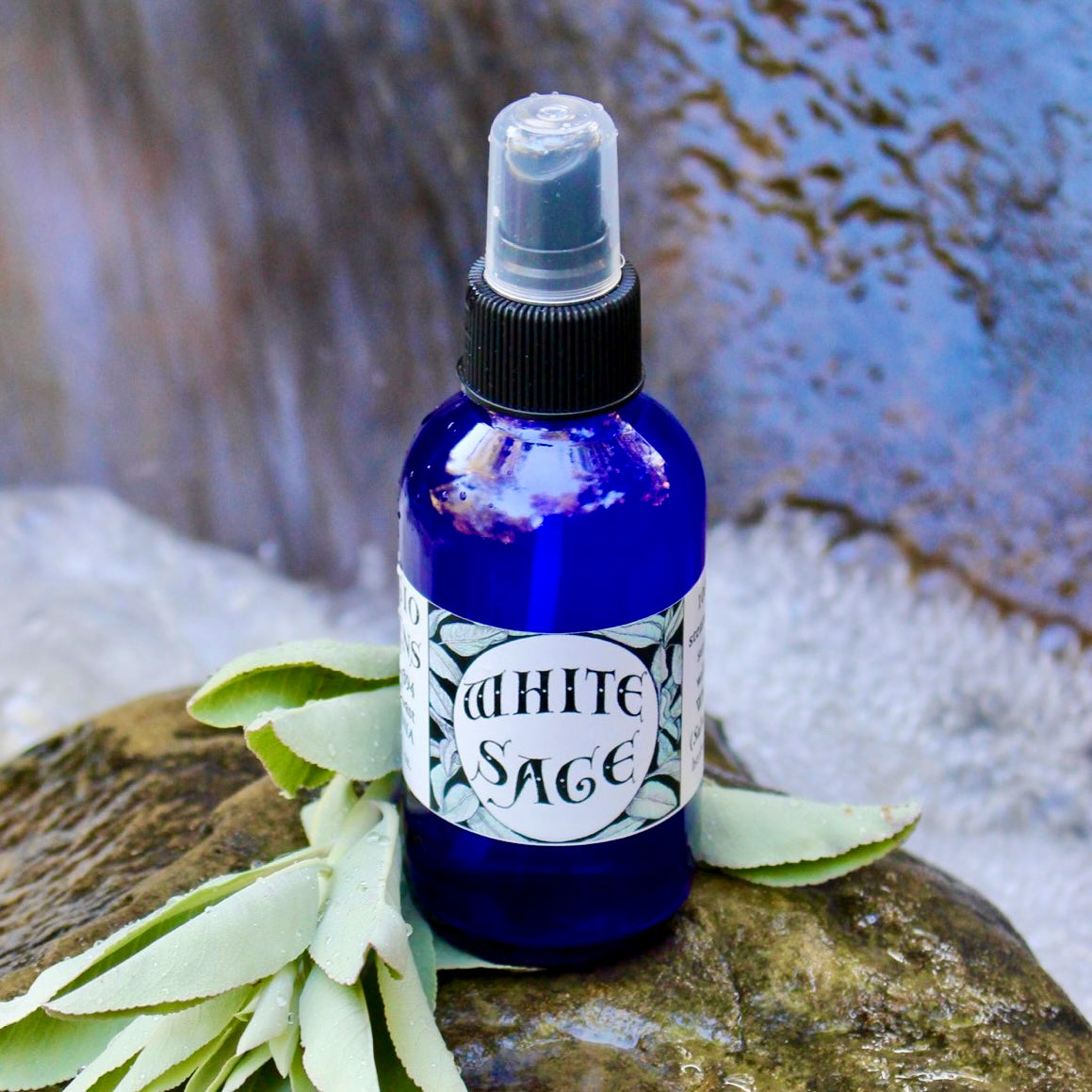 Photograph of a 4 oz cobalt blue glass spray bottle filled with White Sage hydrosol on rock withwhite sage leaves in a creek